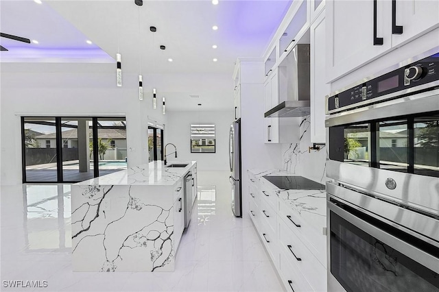 kitchen with a large island with sink, a sink, stainless steel appliances, white cabinets, and wall chimney range hood