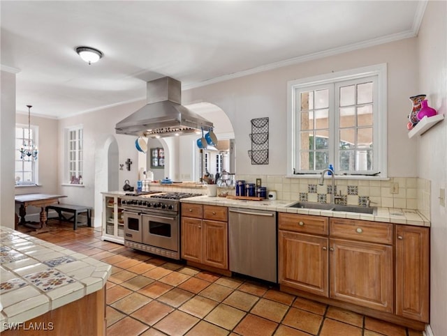 kitchen with tile counters, island exhaust hood, arched walkways, stainless steel appliances, and a sink