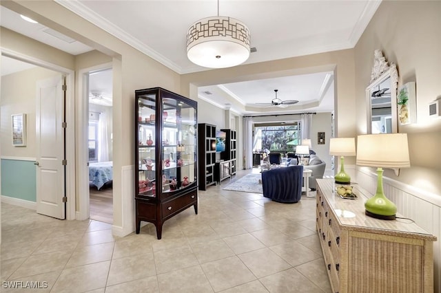 corridor featuring crown molding, light tile patterned floors, a wainscoted wall, and a raised ceiling