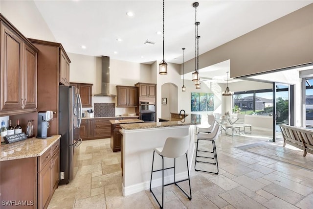 kitchen featuring stone tile floors, a spacious island, appliances with stainless steel finishes, a kitchen breakfast bar, and wall chimney exhaust hood