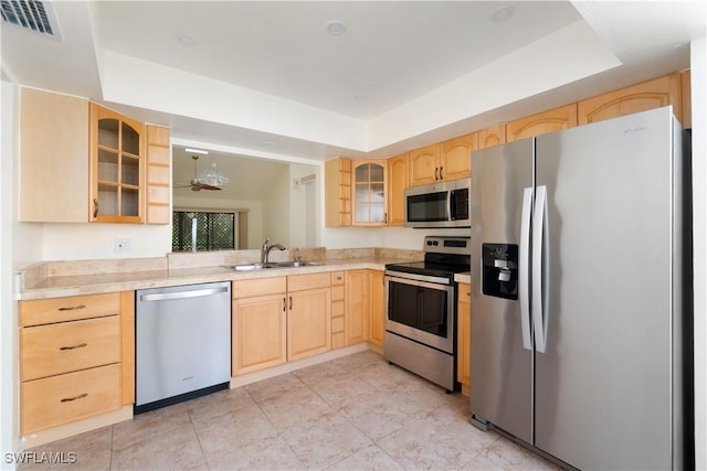 kitchen featuring glass insert cabinets, light brown cabinetry, stainless steel appliances, a raised ceiling, and a sink