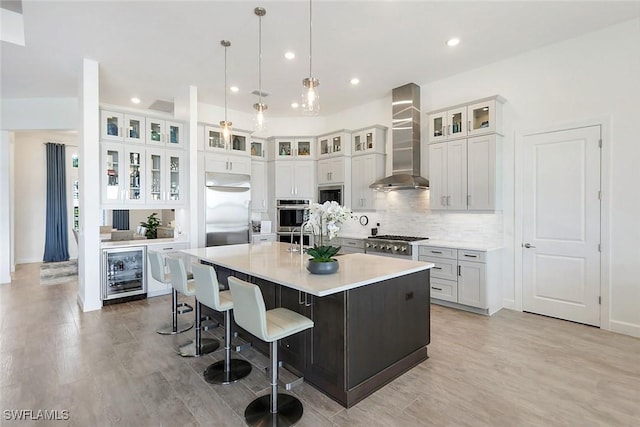 kitchen featuring beverage cooler, built in appliances, a center island with sink, decorative backsplash, and wall chimney exhaust hood