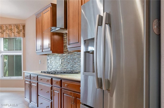 kitchen featuring tasteful backsplash, light tile patterned flooring, brown cabinetry, stainless steel appliances, and wall chimney exhaust hood