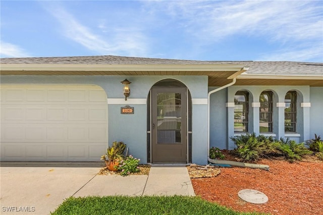 doorway to property featuring a shingled roof, a garage, driveway, and stucco siding