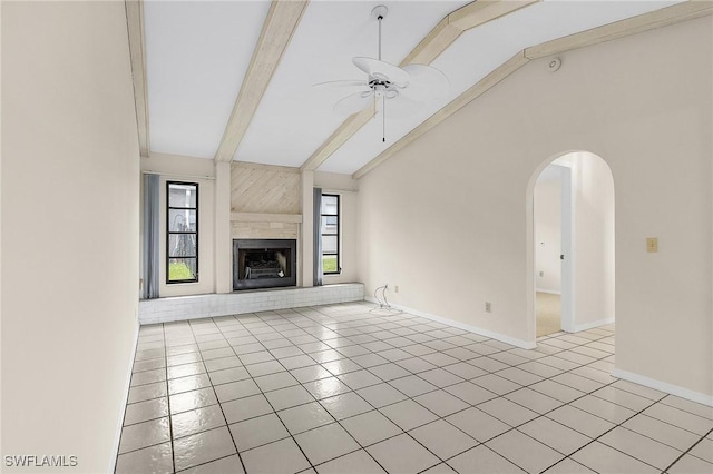 unfurnished living room featuring a fireplace with raised hearth, lofted ceiling with beams, arched walkways, light tile patterned floors, and ceiling fan