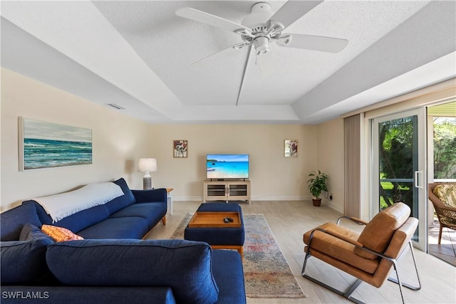 living room with a raised ceiling, a ceiling fan, a textured ceiling, light wood finished floors, and baseboards