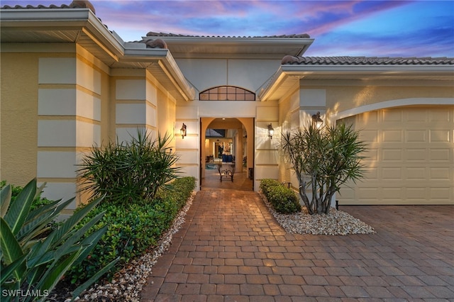 exterior entry at dusk featuring stucco siding, decorative driveway, a garage, and a tiled roof