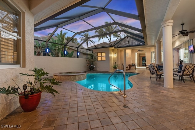 pool at dusk featuring glass enclosure, a pool with connected hot tub, ceiling fan, and a patio area