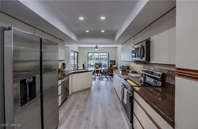 kitchen with dark stone countertops, stainless steel appliances, decorative backsplash, a raised ceiling, and light wood-type flooring