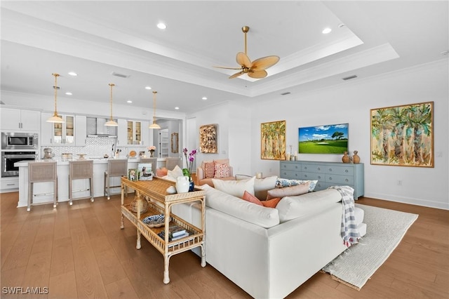 living area featuring visible vents, a tray ceiling, ornamental molding, light wood-style flooring, and a ceiling fan