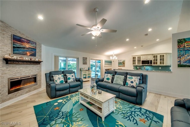 living area featuring visible vents, light wood finished floors, lofted ceiling, a stone fireplace, and ceiling fan with notable chandelier
