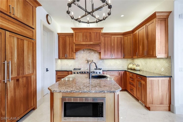 kitchen with light stone counters, a center island with sink, decorative backsplash, stainless steel microwave, and brown cabinets