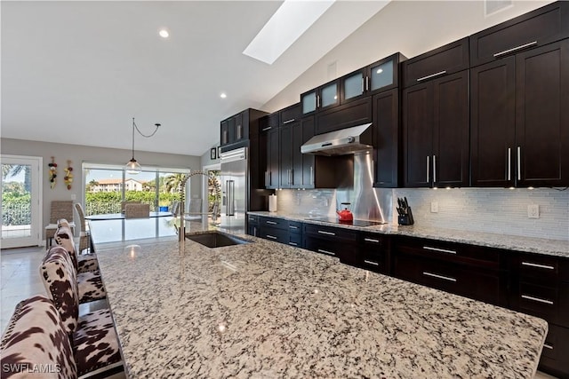 kitchen with under cabinet range hood, light stone countertops, backsplash, and a kitchen island with sink