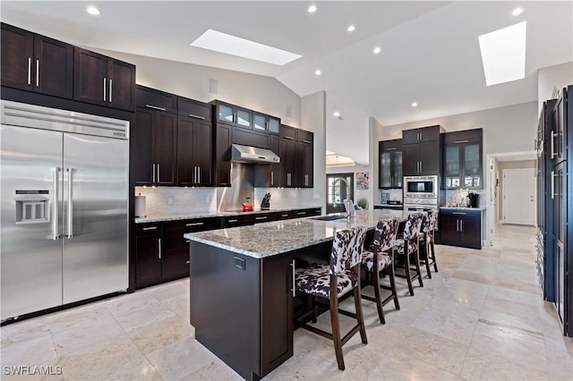 kitchen featuring an island with sink, glass insert cabinets, under cabinet range hood, built in appliances, and lofted ceiling with skylight