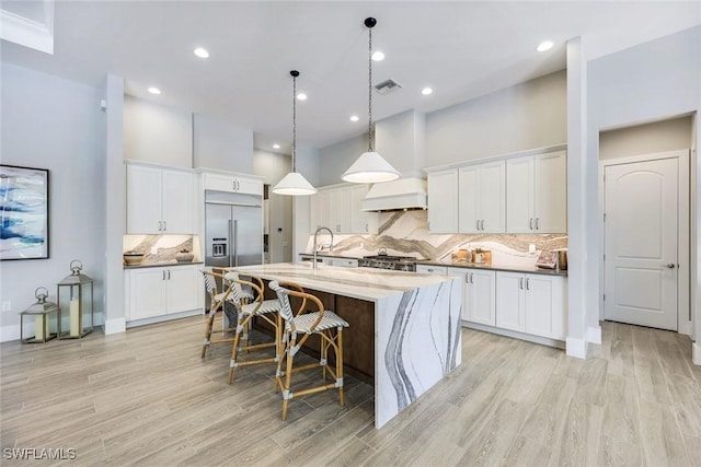 kitchen featuring premium range hood, a kitchen bar, an island with sink, white cabinetry, and built in fridge