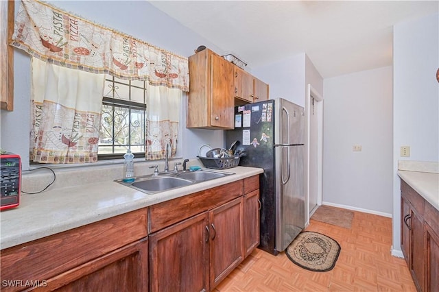 kitchen featuring baseboards, light countertops, freestanding refrigerator, brown cabinetry, and a sink