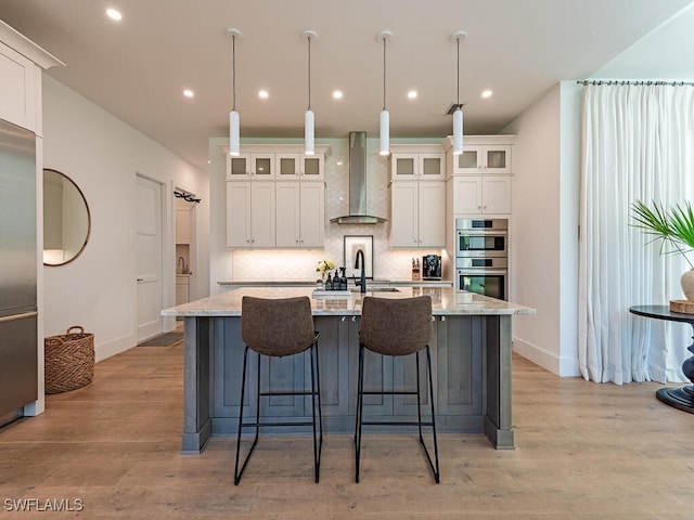 kitchen with light wood-type flooring, an island with sink, backsplash, appliances with stainless steel finishes, and wall chimney range hood