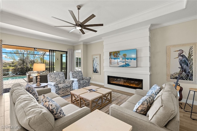 living area with a glass covered fireplace, a raised ceiling, wood finished floors, and a ceiling fan