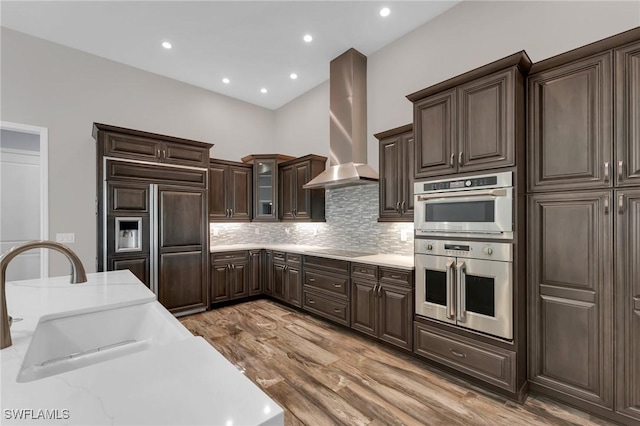 kitchen featuring wall chimney range hood, double oven, decorative backsplash, black electric cooktop, and a sink