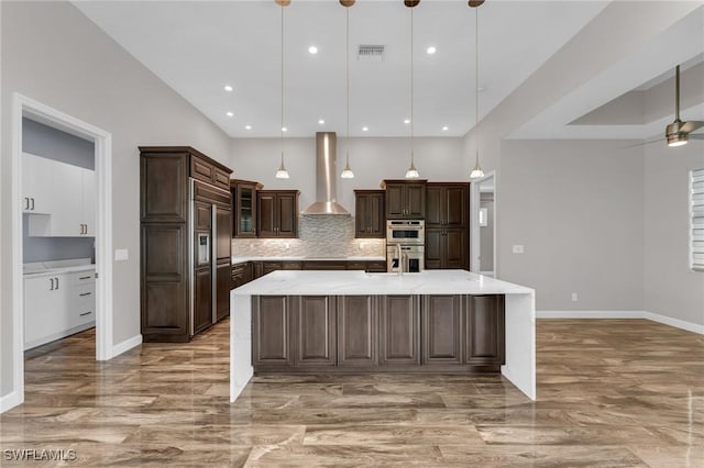 kitchen with a large island, backsplash, double oven, wall chimney exhaust hood, and dark brown cabinets