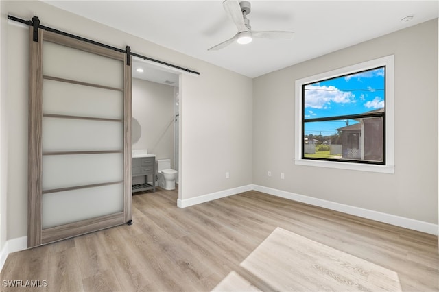 unfurnished bedroom with light wood finished floors, connected bathroom, baseboards, a barn door, and a ceiling fan