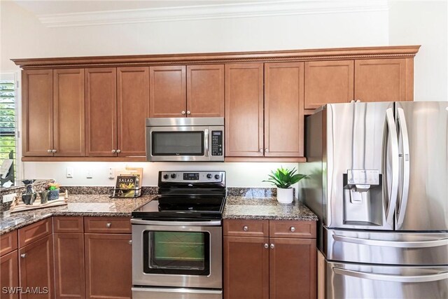 kitchen featuring brown cabinets, stainless steel appliances, crown molding, and dark stone counters