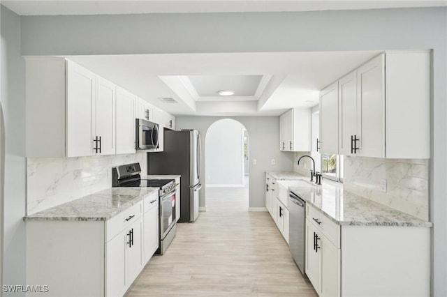 kitchen with light stone countertops, arched walkways, stainless steel appliances, white cabinets, and a raised ceiling