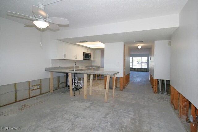 kitchen featuring a ceiling fan, a peninsula, white cabinets, stainless steel microwave, and unfinished concrete flooring