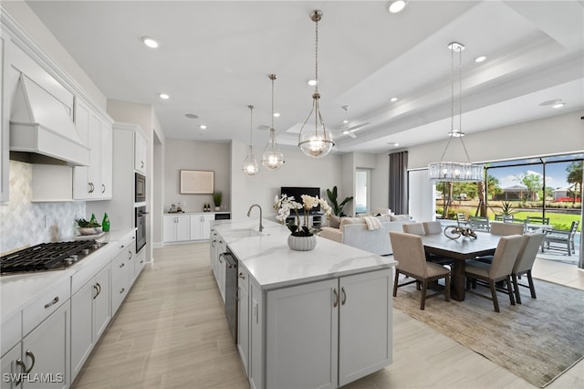 kitchen with tasteful backsplash, custom range hood, appliances with stainless steel finishes, an inviting chandelier, and a kitchen island with sink