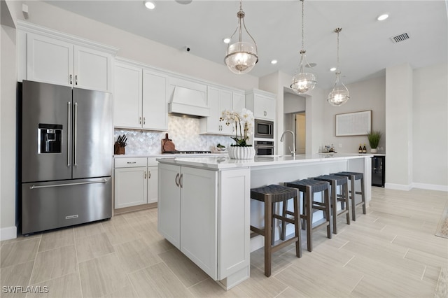 kitchen featuring custom exhaust hood, an island with sink, white cabinets, appliances with stainless steel finishes, and tasteful backsplash