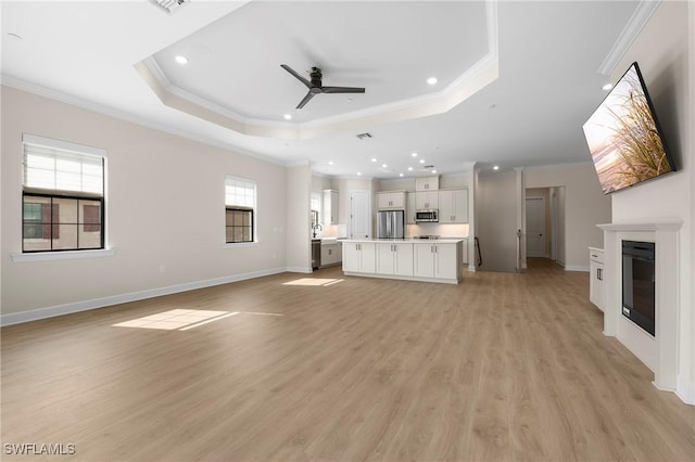 unfurnished living room with light wood-style flooring, crown molding, ceiling fan, a raised ceiling, and a glass covered fireplace