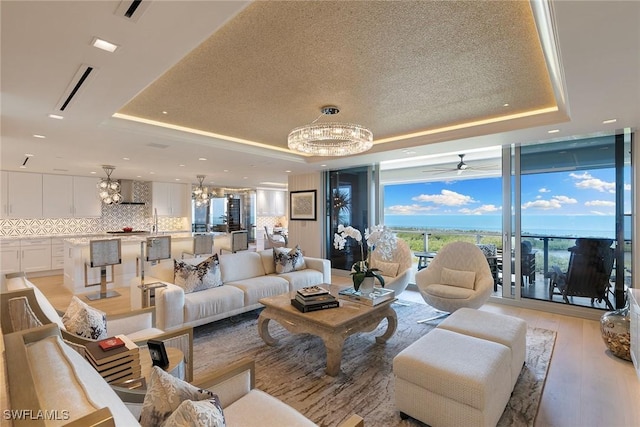 living room with ceiling fan with notable chandelier, a tray ceiling, light wood-style floors, and expansive windows