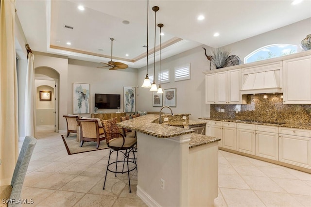 kitchen featuring a kitchen bar, backsplash, a raised ceiling, arched walkways, and custom exhaust hood