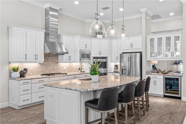 kitchen with wine cooler, wall chimney exhaust hood, appliances with stainless steel finishes, light wood-type flooring, and a center island with sink