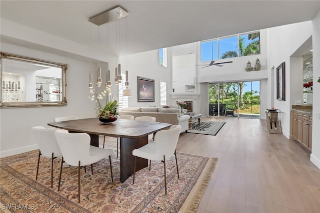 dining room featuring baseboards, a high ceiling, ceiling fan, a fireplace, and light wood-style flooring
