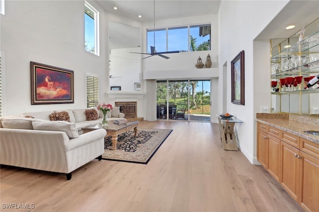 living area with a towering ceiling, recessed lighting, a fireplace with raised hearth, ceiling fan, and light wood finished floors
