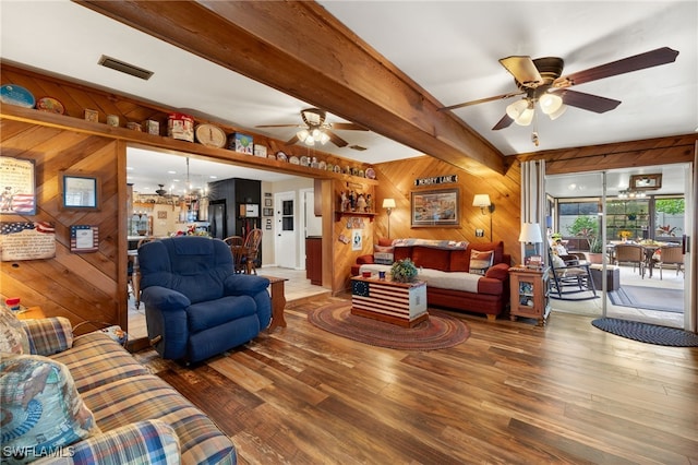 living room with ceiling fan with notable chandelier, beam ceiling, wooden walls, visible vents, and wood finished floors