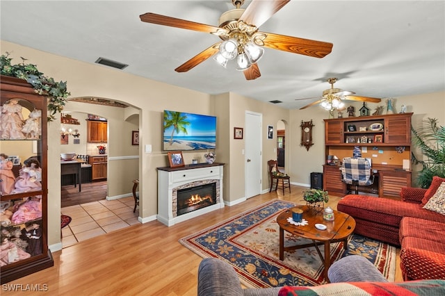 living area featuring a ceiling fan, a glass covered fireplace, light wood-type flooring, visible vents, and arched walkways