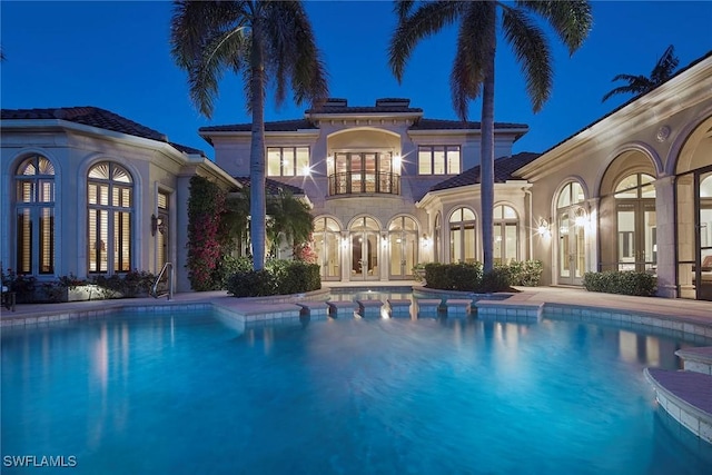 back of house at night with a balcony, french doors, stucco siding, and an outdoor pool