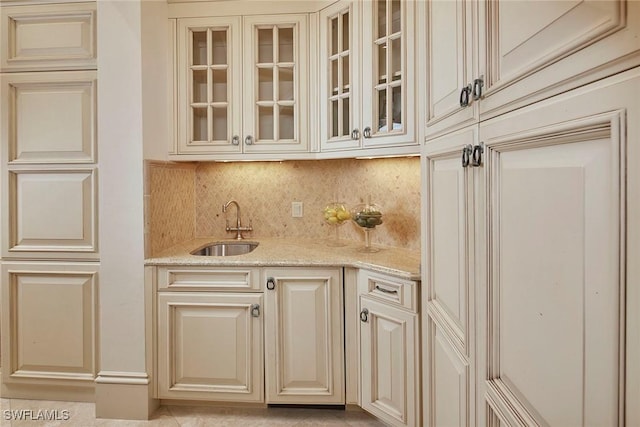 kitchen featuring cream cabinetry, glass insert cabinets, a sink, light stone counters, and backsplash