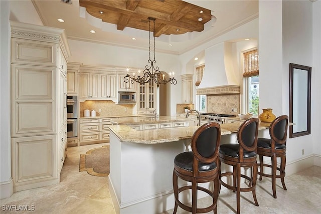 kitchen with coffered ceiling, stainless steel appliances, cream cabinets, light stone countertops, and custom exhaust hood