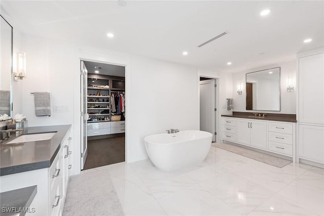 full bath featuring two vanities, recessed lighting, a soaking tub, a walk in closet, and marble finish floors