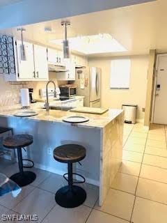 kitchen with a peninsula, stainless steel microwave, under cabinet range hood, light tile patterned floors, and freestanding refrigerator