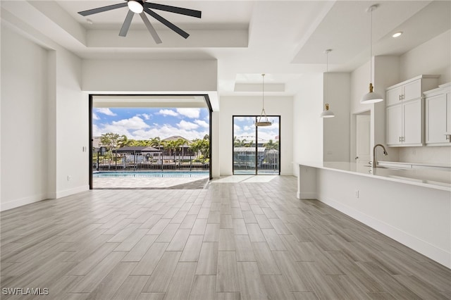 unfurnished living room featuring light wood-style flooring, a tray ceiling, a sink, baseboards, and ceiling fan