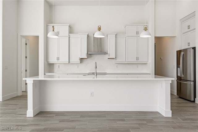 kitchen with wall chimney exhaust hood, a kitchen island with sink, light wood finished floors, stainless steel fridge with ice dispenser, and light countertops