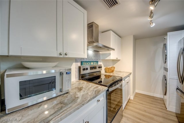 kitchen featuring stainless steel electric range oven, wall chimney exhaust hood, stacked washer / drying machine, white microwave, and light wood finished floors