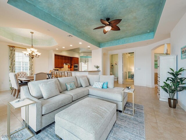 living area featuring light tile patterned floors, a tray ceiling, baseboards, arched walkways, and ceiling fan with notable chandelier
