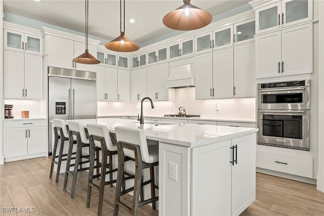 kitchen with light countertops, appliances with stainless steel finishes, light wood finished floors, a center island with sink, and custom range hood