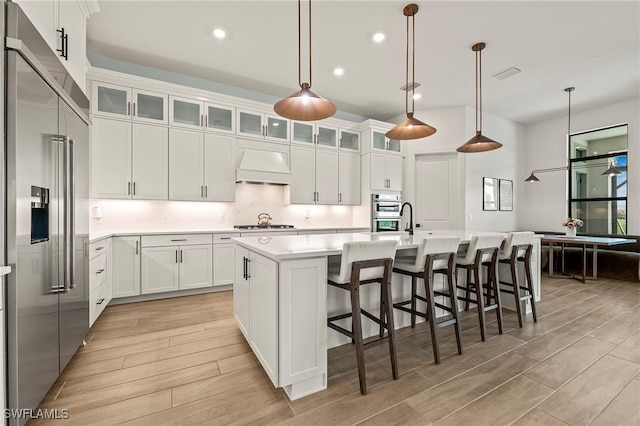 kitchen with built in refrigerator, an island with sink, premium range hood, light countertops, and white cabinets