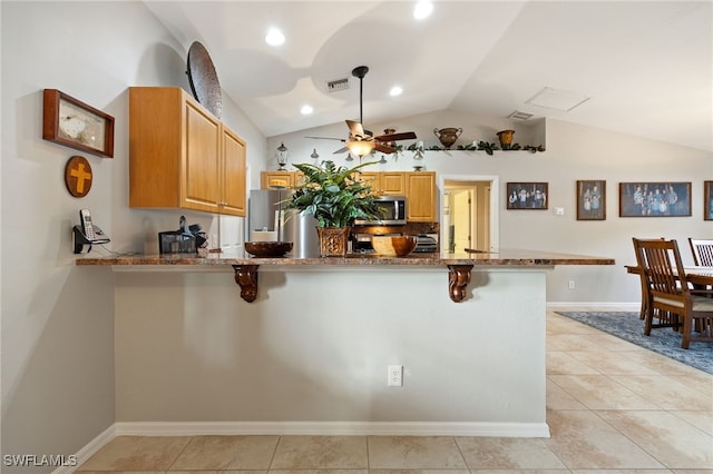 kitchen with a peninsula, ceiling fan, appliances with stainless steel finishes, dark stone countertops, and a breakfast bar area
